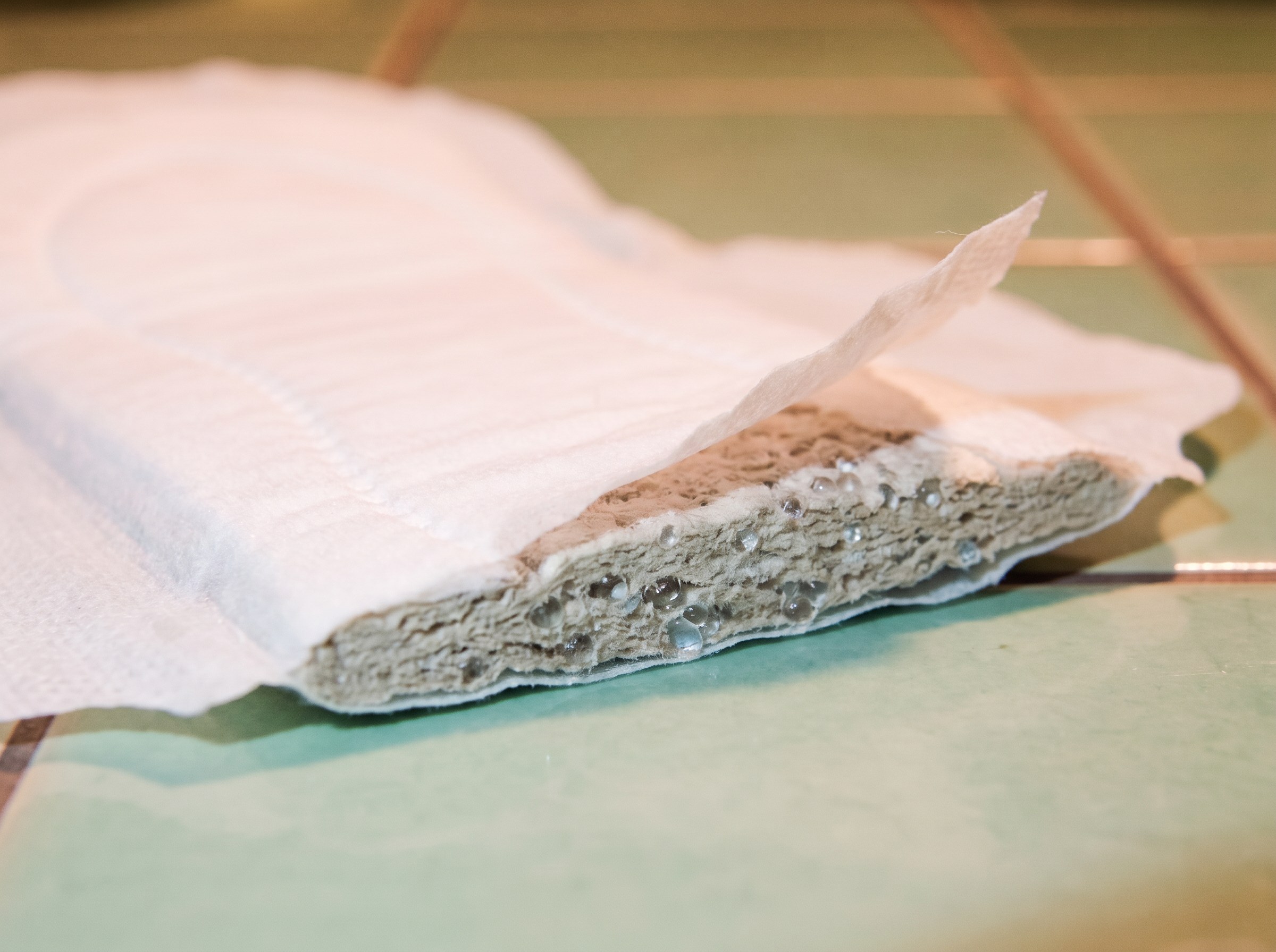 A generic pad cut open: a thin cotton top sheet lifted away from a dense grey synthetic core